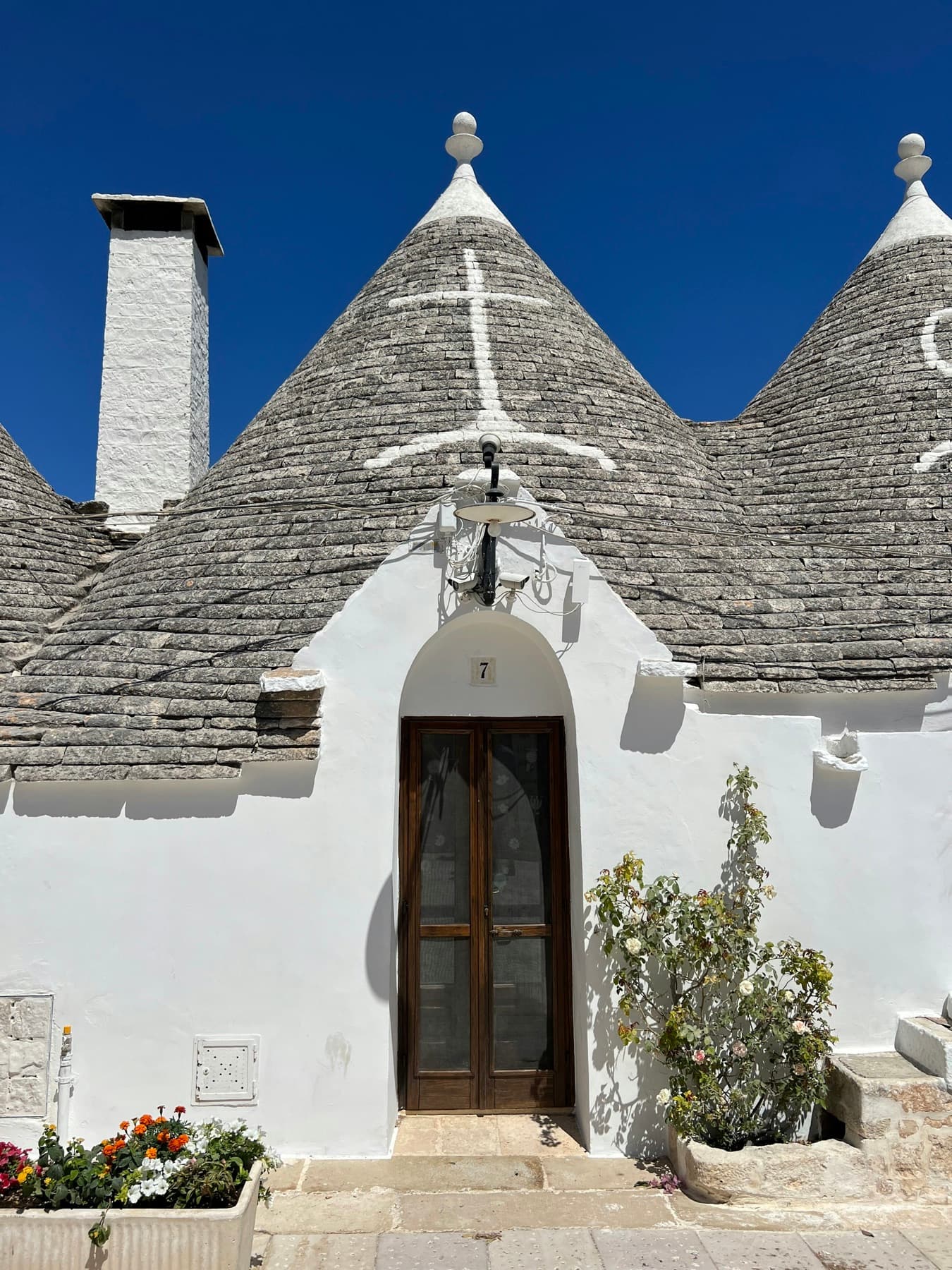 A trullo with three conical stone roofs, a white wall and a wooden door under a deep blue sky, Alberobello